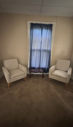 A room with beige walls, a large window with sheer curtains, and two beige armchairs with wooden legs.