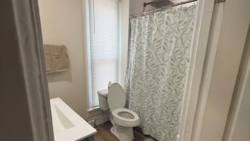 Une salle de bain avec des toilettes, un rideau de douche et une fenêtre avec des stores.