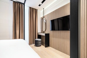 Una habitación de hotel moderna con una cama grande, un televisor de pantalla plana montado en una pared de paneles de madera, un taburete redondo pequeño y una mesita de noche.