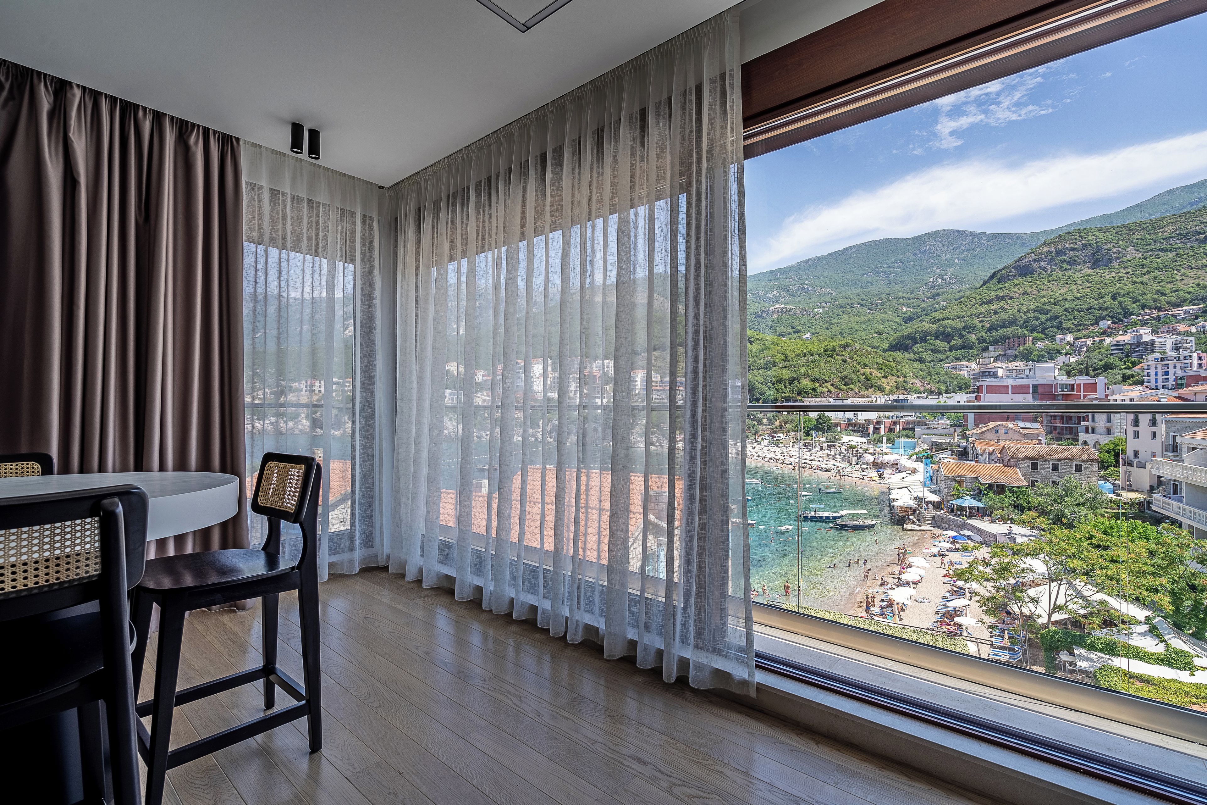 A room with a dining table and chairs, a large window with sheer curtains, and a view of a coastal town.
