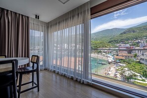 A room with a dining table and chairs, a large window with sheer curtains, and a view of a coastal town.