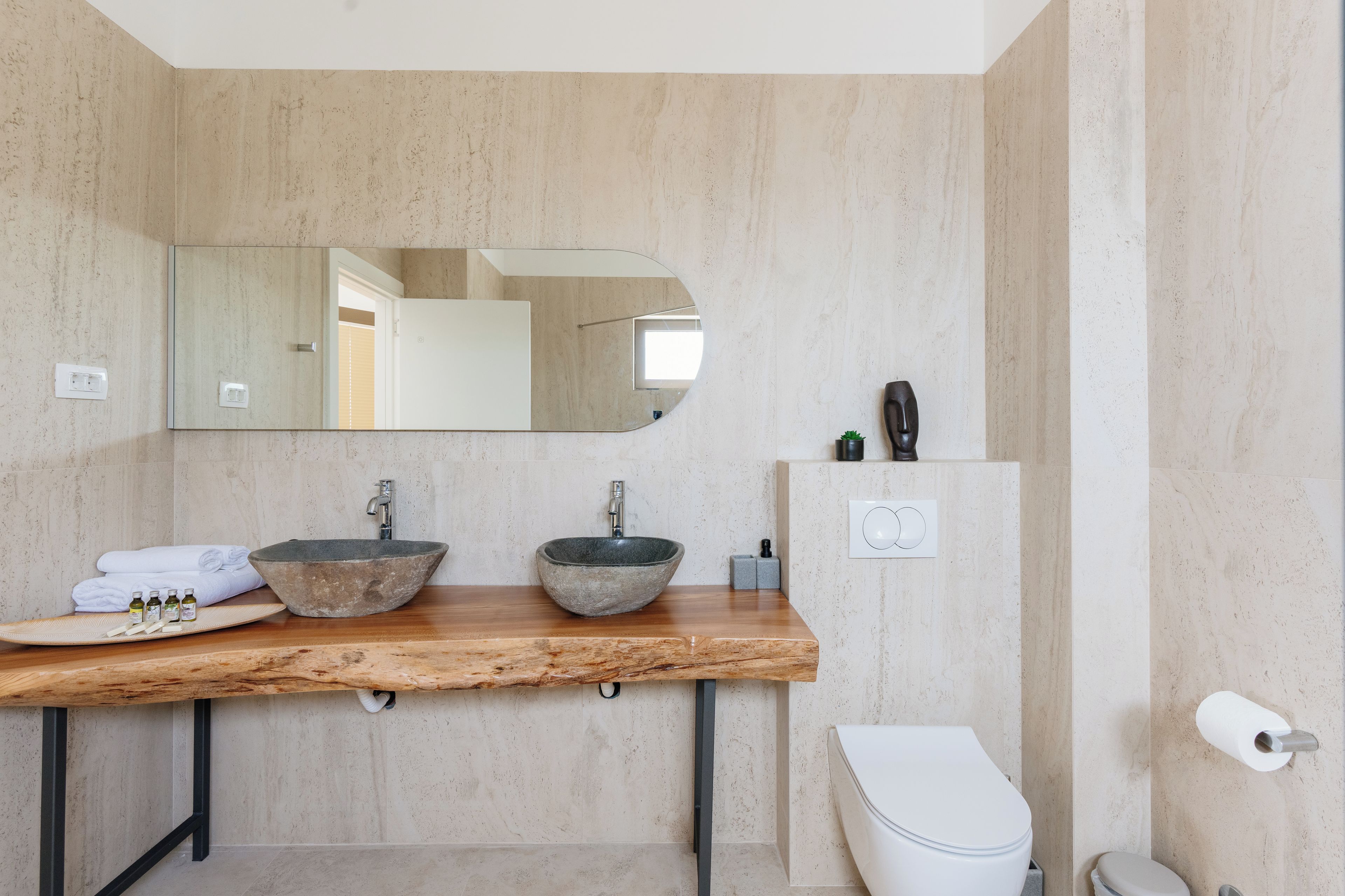 A bathroom with a wooden vanity, two stone sinks, a mirror, a toilet, and a small shelf with toiletries.
