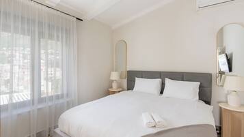 A hotel room with a bed, two bedside tables with lamps, a large window with sheer curtains, and a city view.
