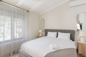A hotel room with a bed, two bedside tables with lamps, a large window with sheer curtains, and a city view.