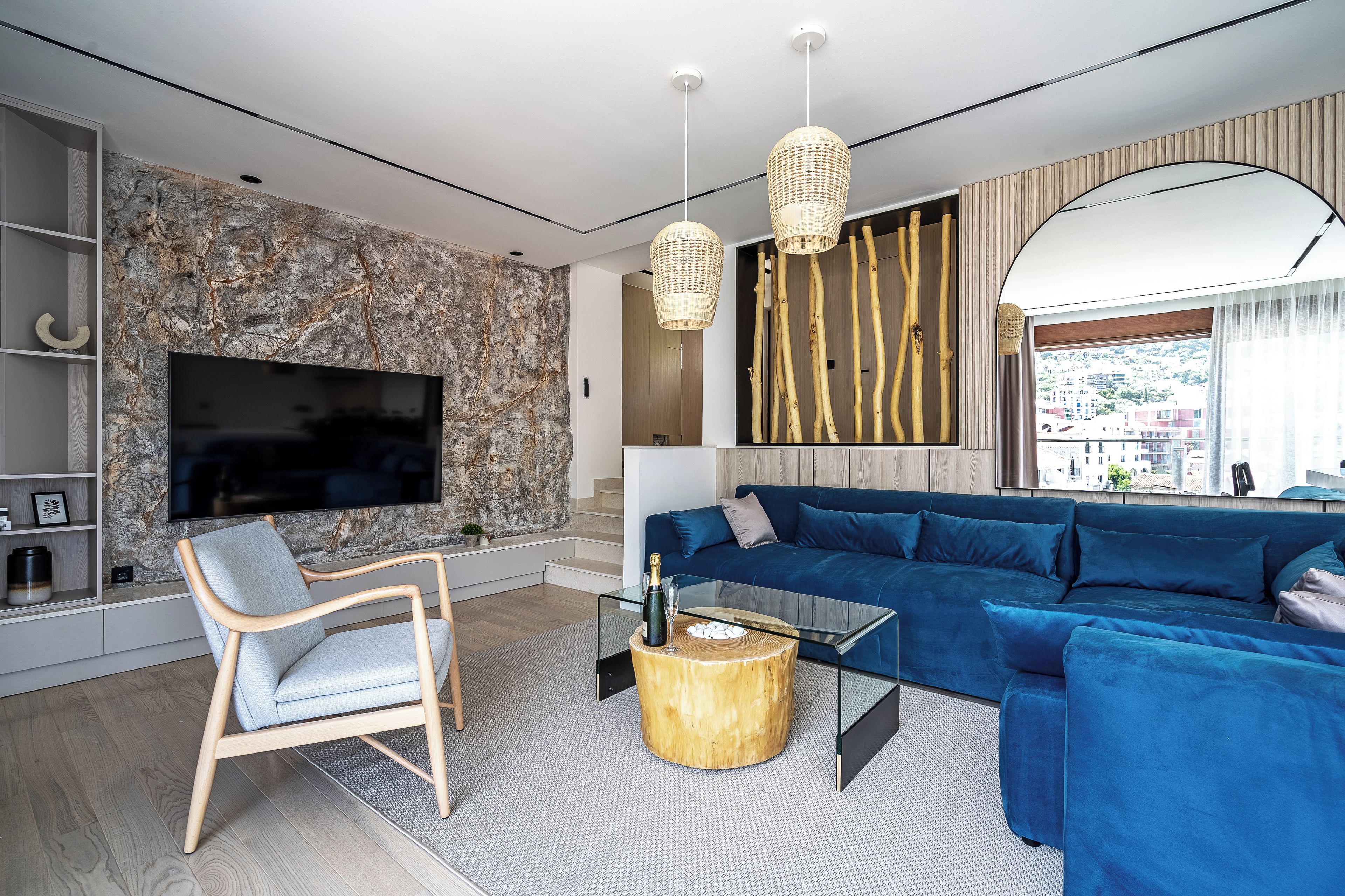 Ein modernes Wohnzimmer mit einer Wand aus Stein, einem großen Flachbildfernseher, einem blauen Sofa, einem Holztisch und einer Pendelleuchte aus Rattan.