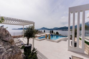 Zona de piscina con sillones de descanso y una pérgola con vistas a un cuerpo de agua y montañas.