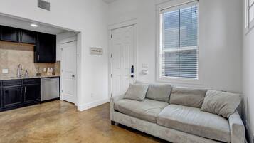 Et moderne køkken med mørke skabe, en bordplade og en opvaskemaskine. Der er en beige sofa i en stue med et vindue og persienner.