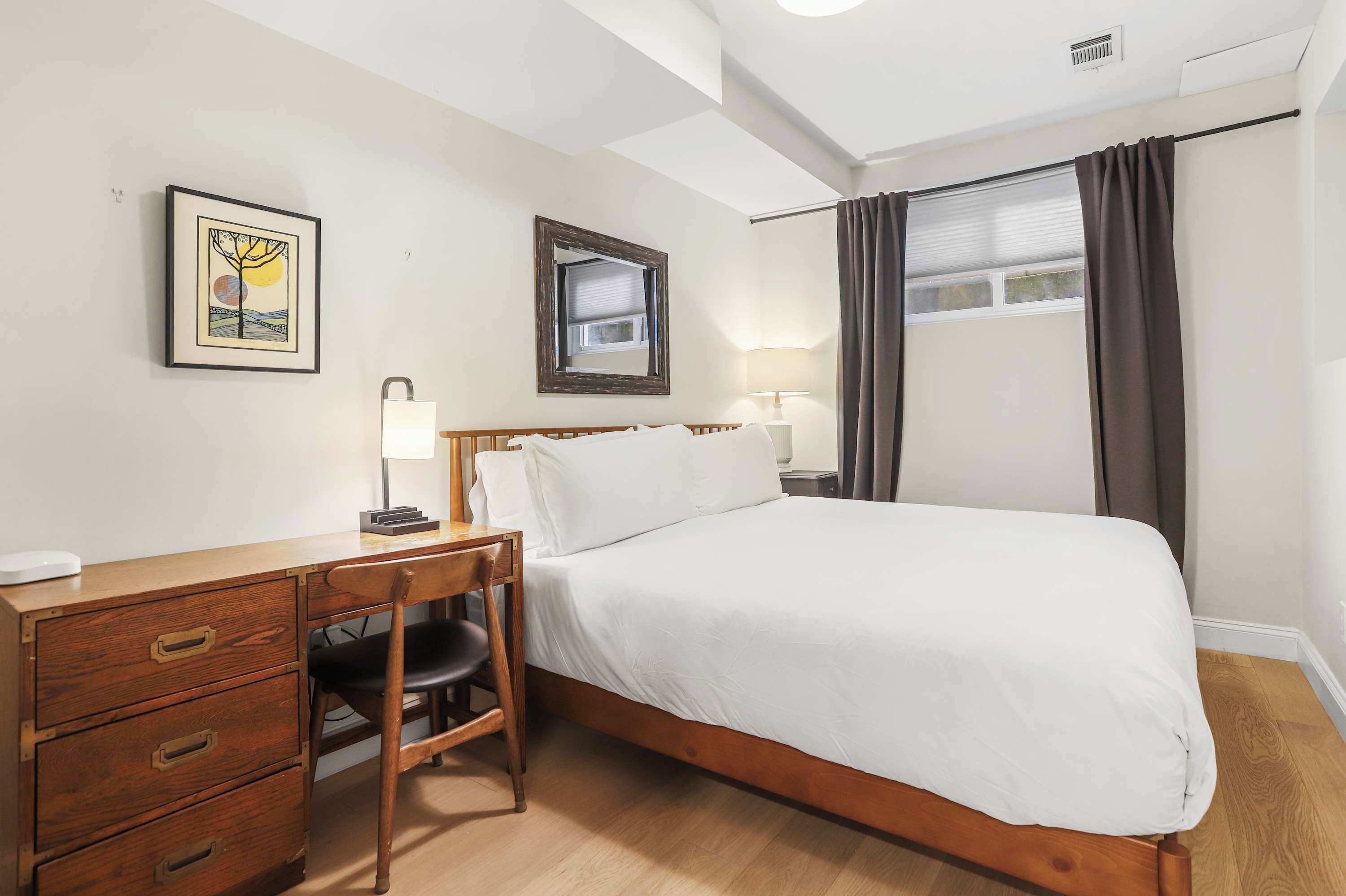 Habitación de hotel con cama de madera, un escritorio con cajones, una silla, una mesita de noche con lámpara, un cuadro en la pared y una ventana con cortinas.