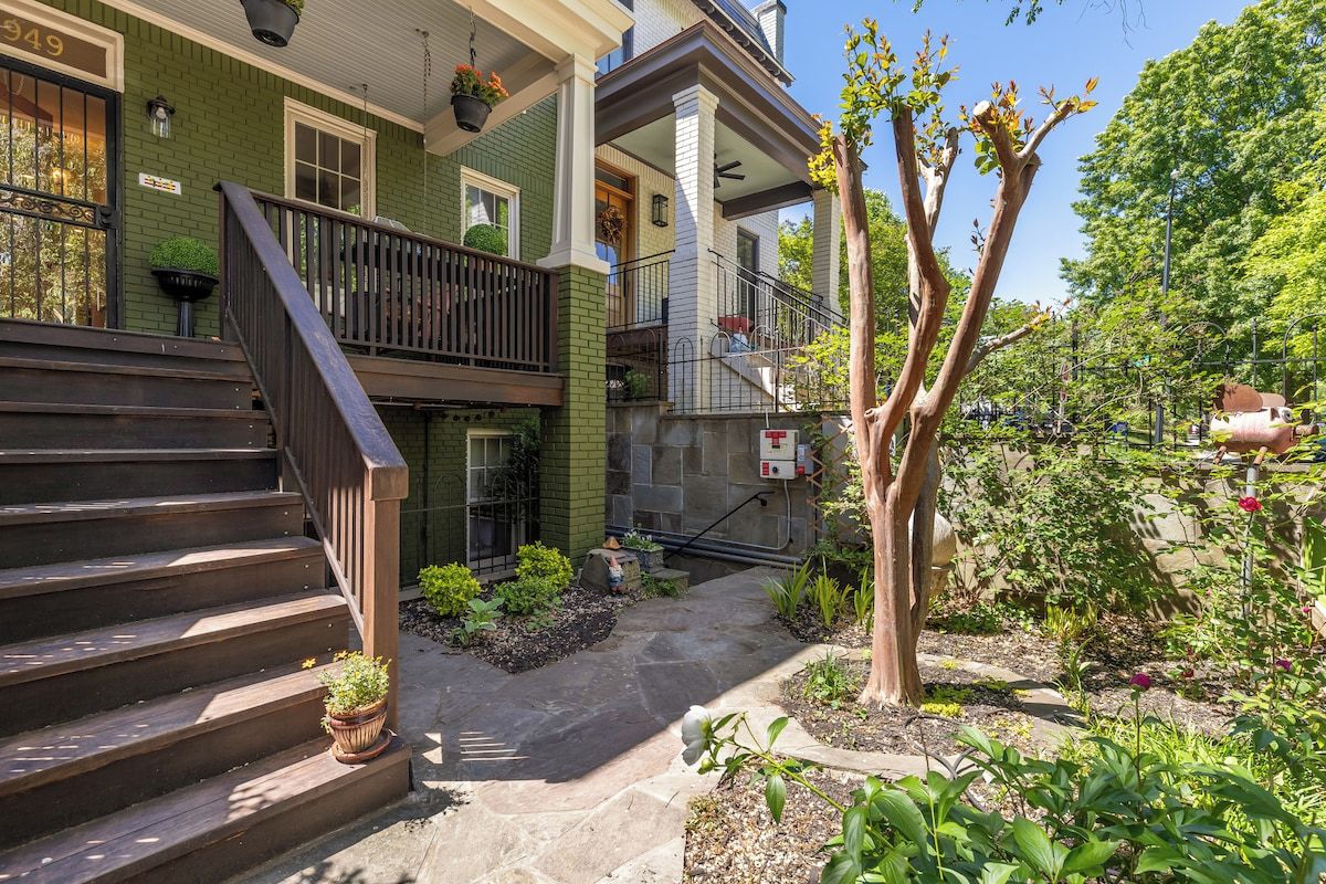 Une maison à deux étages avec un porche à l’avant, un escalier et un jardin avec un arbre.