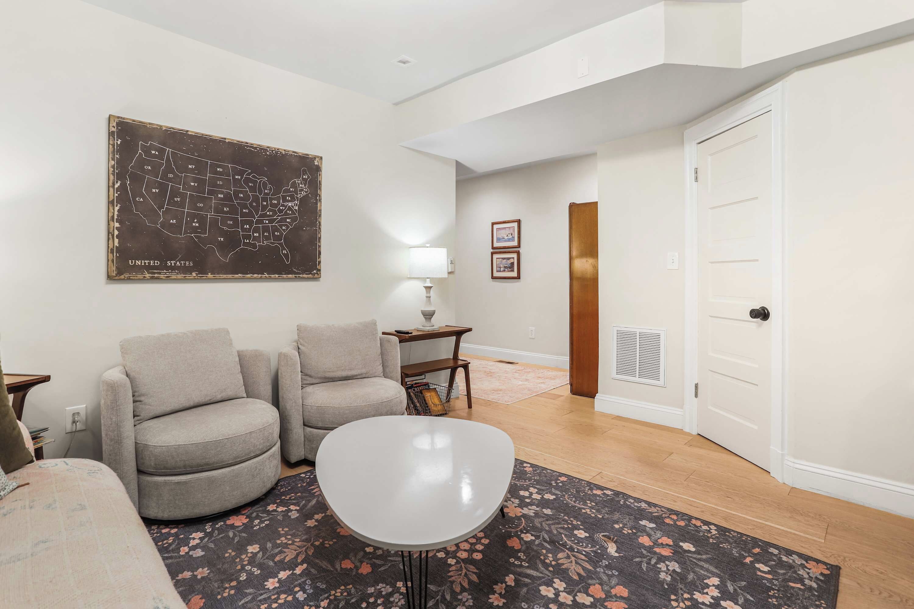 A living room with two armchairs, a coffee table, a map on the wall, a lamp, and a door leading to another room.