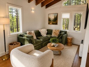 A cozy living room with a green sofa, a white armchair, and a round wooden coffee table. There is a painting on the wall, a lamp, and a window with a view of trees. - Charming Updated Tahoe Cabin Near Heavenly | Forest Views | Two Decks (South Lake Tahoe)