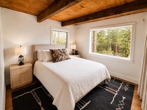 A bedroom with a large bed, two bedside tables, a window with a forest view, and a wooden ceiling. - Charming Updated Tahoe Cabin Near Heavenly | Forest Views | Two Decks (South Lake Tahoe)