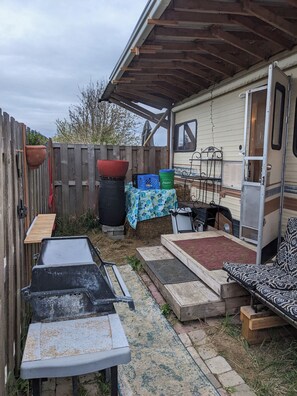 A covered outdoor area with a wooden table, a grill, a seating area with a patterned cushion, and a covered RV. - Charming 2-bedroom recreational vehicle in enjoyable Sequim location (Sequim)