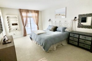 Ein Schlafzimmer mit Bett, Nachttischen, Schreibtisch, Stuhl, einem Fenster mit Vorhängen und einem an der Wand montierten Fernseher.