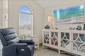 A modern living room with a leather recliner, a television mounted on the wall, a decorative cabinet with glass doors, and a window with blinds. - Beautiful Beach home in a gated neighborhood located near everything! (Panama City Beach)