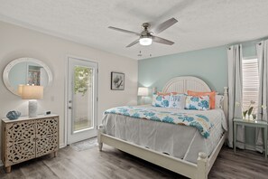 A bedroom with a bed, a ceiling fan, a round mirror, a side table, and a door. - Beautiful Beach home in a gated neighborhood located near everything! (Panama City Beach)