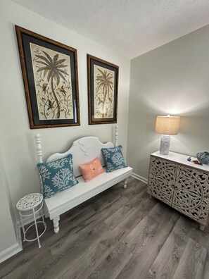 Quarto com um banco branco, duas gravuras botânicas emolduradas, um abajur branco e um armário decorativo.