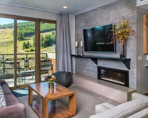 Una sala moderna con chimenea, un televisor de pantalla plana y vistas a un paisaje boscoso a través de amplios ventanales correderas.