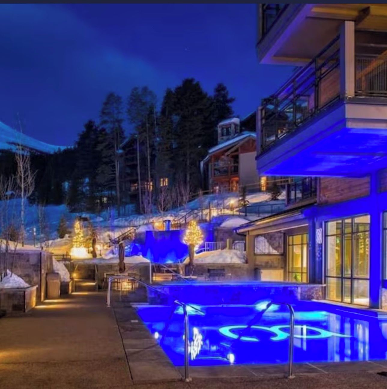 A modern building with a pool area illuminated by blue lights, surrounded by snow-covered ground and trees.