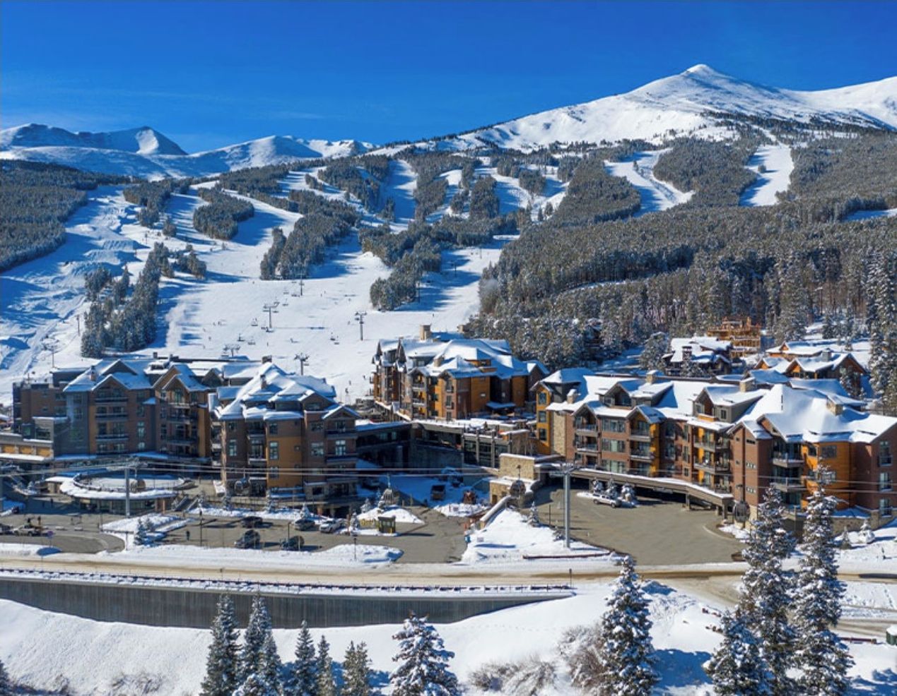 A snow-covered mountain resort with multiple buildings, ski slopes, and a clear blue sky.