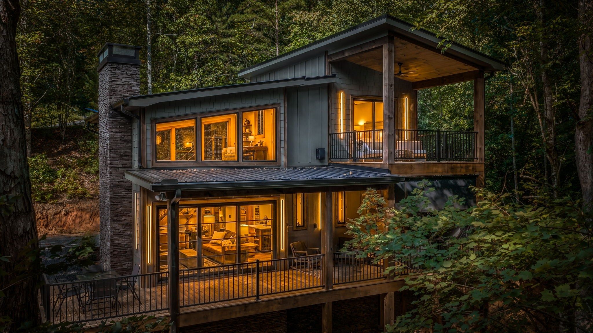 A two-story wooden cabin with large windows and a balcony, surrounded by dense forest.