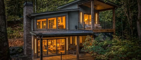 A two-story wooden cabin with large windows and a balcony, surrounded by dense forest.