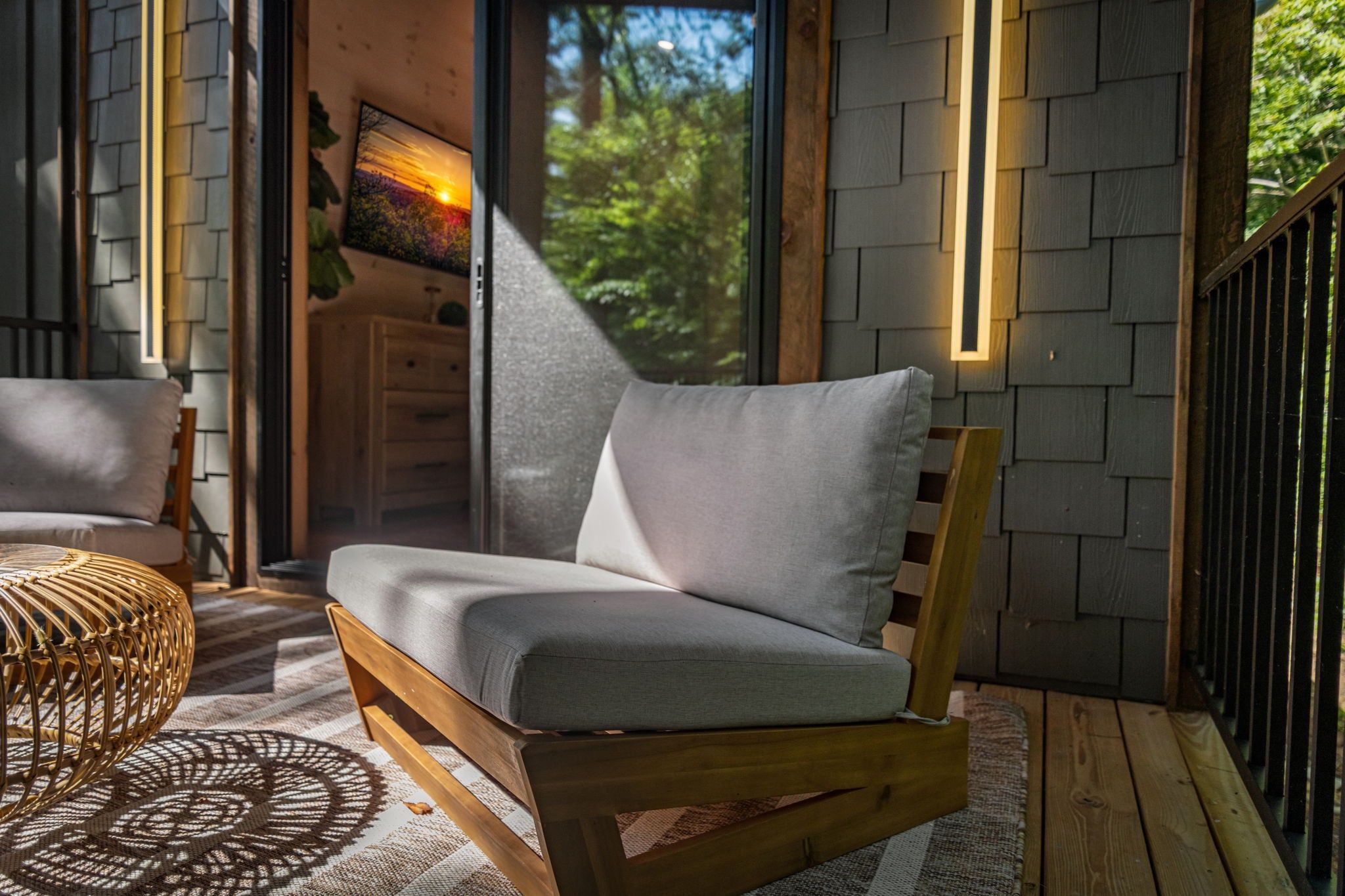 A wooden-framed sofa with grey cushions in a sunlit room with a view of greenery outside.