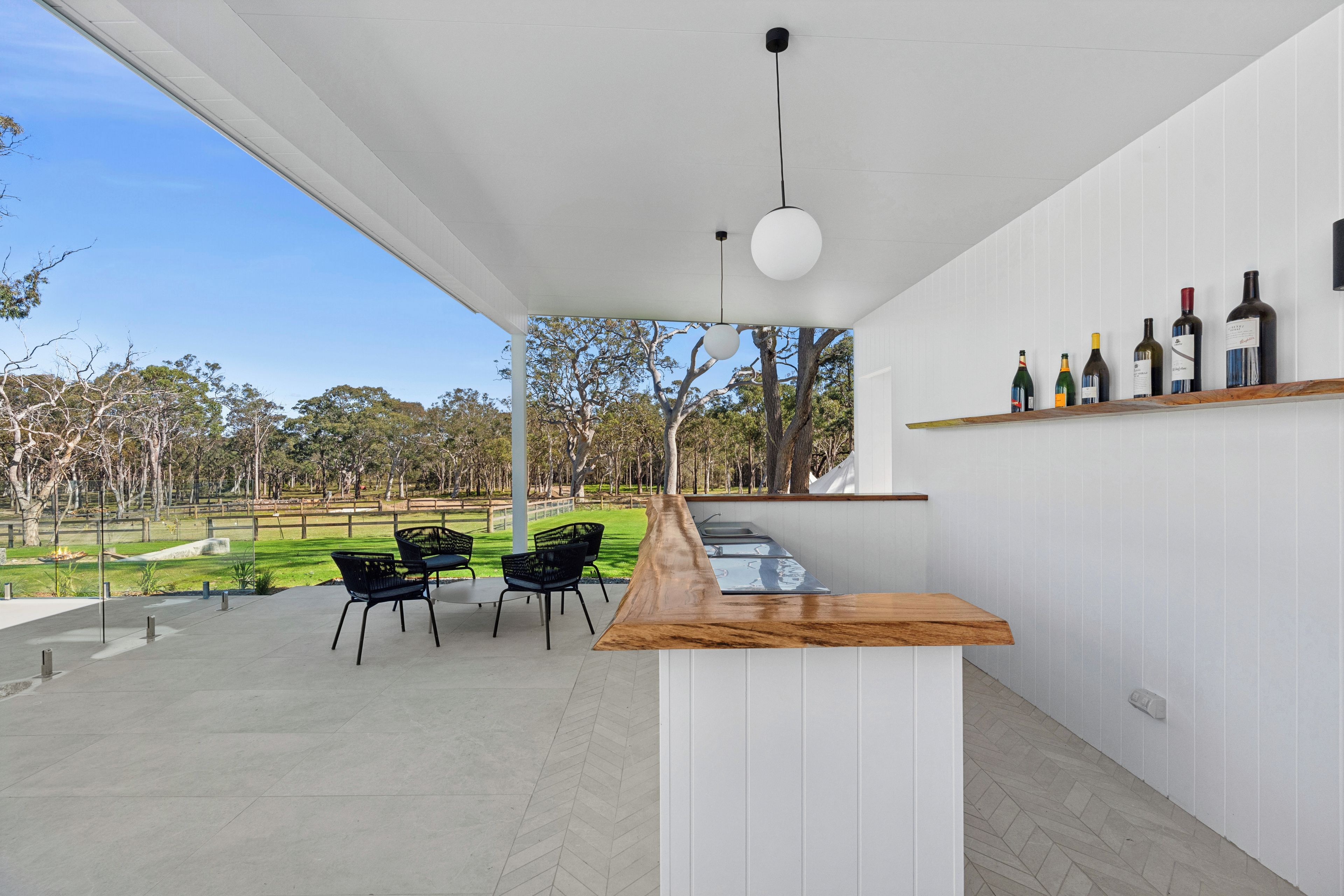 Un espacio al aire libre moderno con barra de madera, sillas negras y una estantería para vinos con botellas.