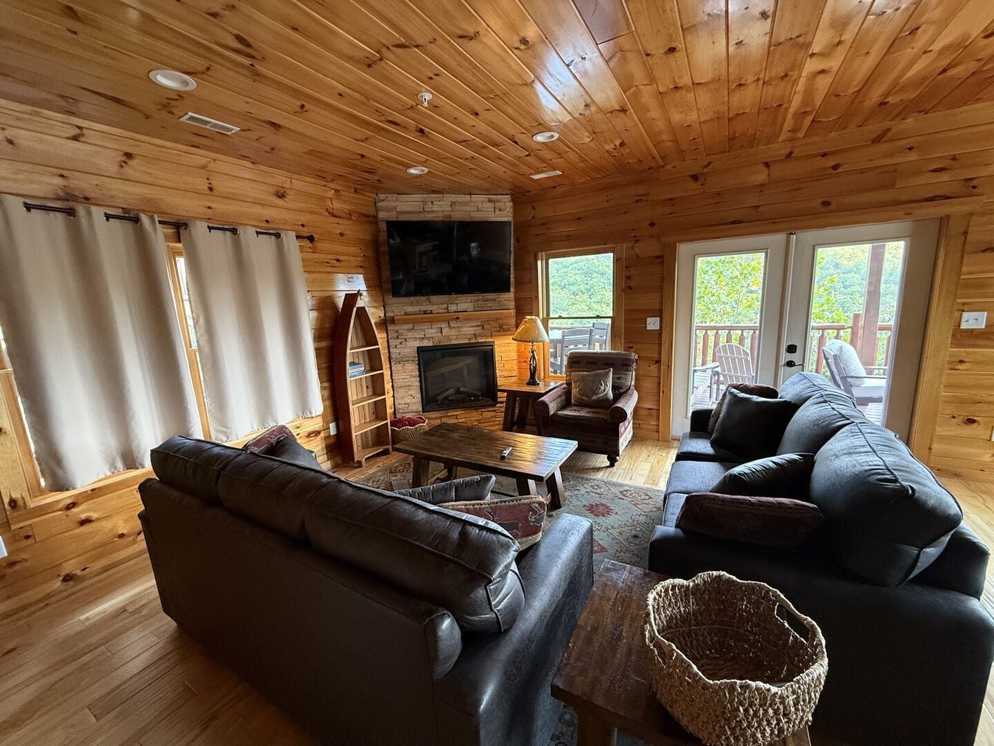 A cozy cabin interior with a fireplace, wooden walls, a television, and a large window.