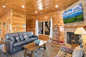 A wooden cabin interior with a leather sofa, a fireplace, and a large window with a view of a mountainous landscape. - Mountain Lookout – Incredible Mt. LeConte Views, Theater, Game Room, Sauna & Hot Tub (Gatlinburg)