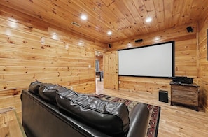 A wooden-paneled room with a black leather sofa, a projector screen, and a speaker. - Mountain Lookout – Incredible Mt. LeConte Views, Theater, Game Room, Sauna & Hot Tub (Gatlinburg)