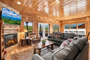 A log cabin interior with a fireplace, seating area, and large windows offering a view of the outdoors. - Mountain Lookout – Incredible Mt. LeConte Views, Theater, Game Room, Sauna & Hot Tub (Gatlinburg)