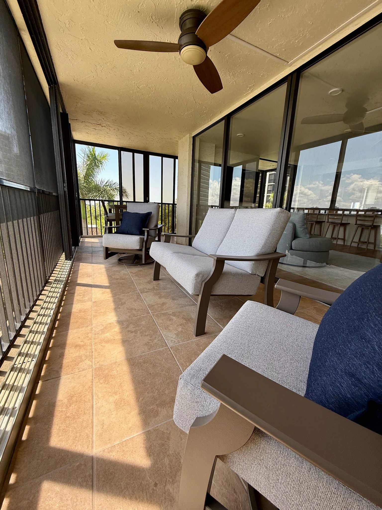 Un balcón soleado con ventilador de techo, zona de estar con sofá y sillones, y vista al cielo y palmeras.