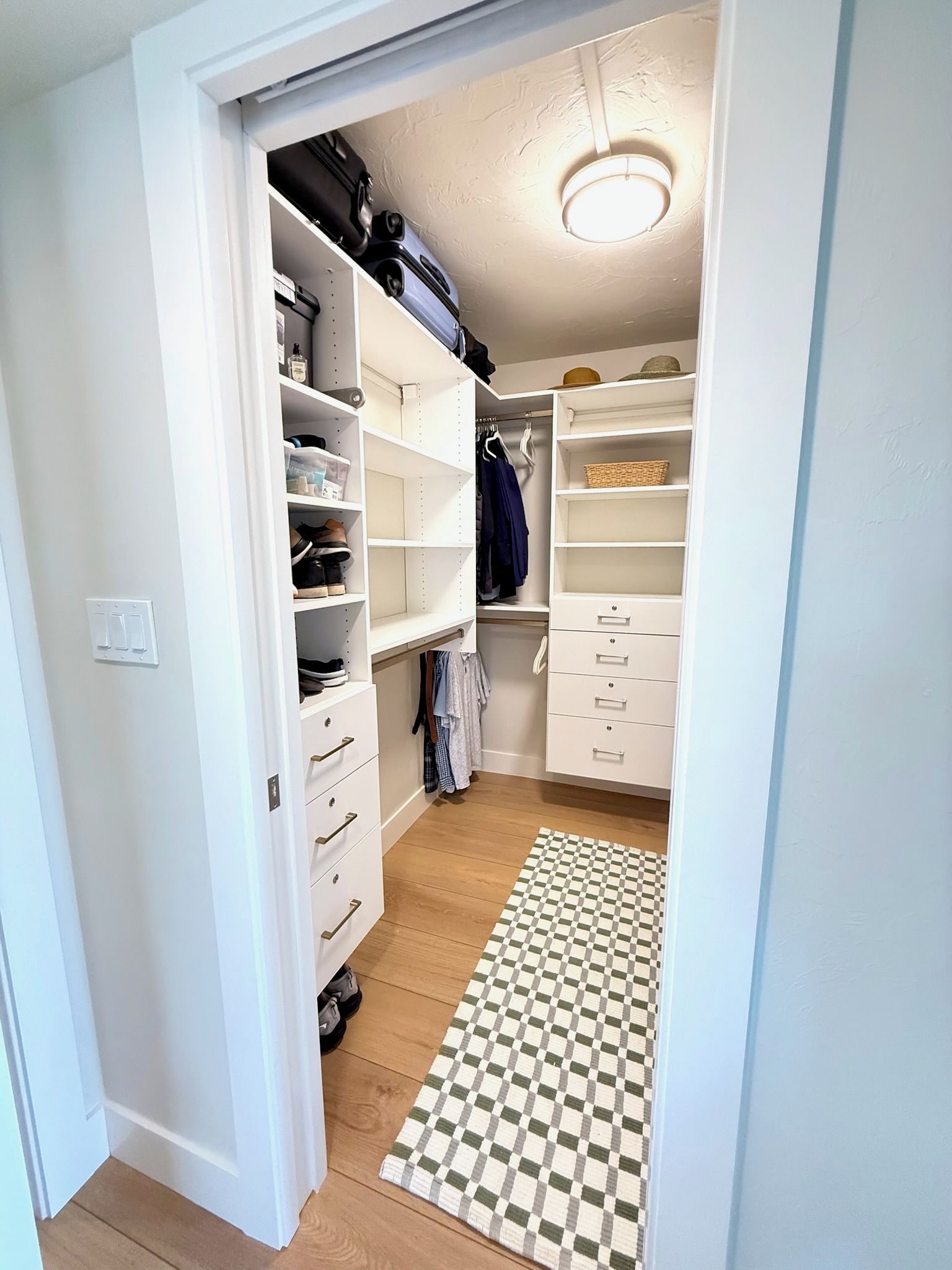 A walk-in closet with shelves, drawers, and hanging space, featuring a checkered rug and a ceiling light.