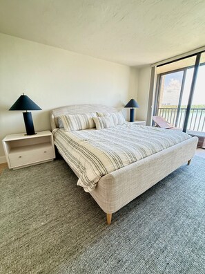 A bedroom with a bed, two bedside tables, and a view of the outdoors through a large window. - Newly Renovated 2 Bedroom Condo across the street from the Beach (Bonita Springs)