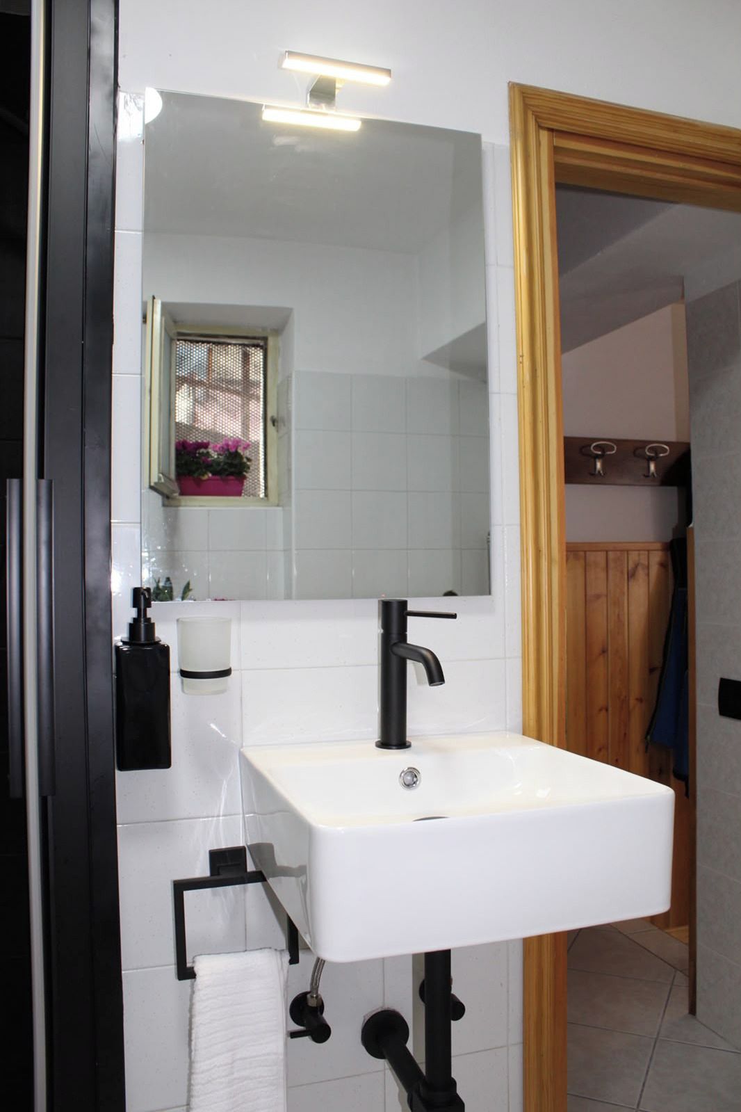 Un baño con un lavamanos blanco, grifo negro y un espejo encima. Hay una ventana con vistas al exterior, y un perchero con una toalla blanca colgada.