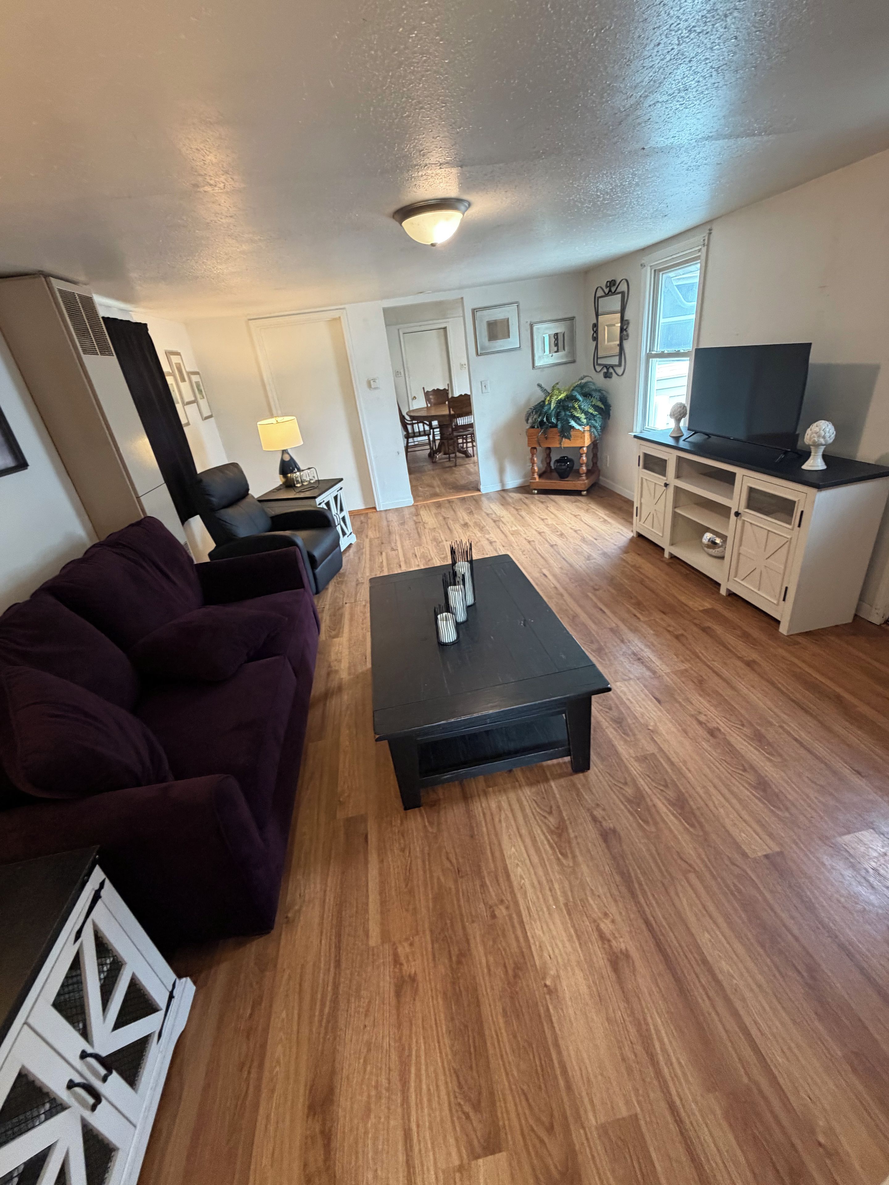 Una sala de estar con una mesa de centro de madera oscura, un sofá morado y un televisor sobre un mueble blanco.