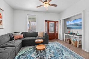 Coastal living room with sectional seating, wall-mounted Smart TV, ceiling fan, patterned area rug, and a bright open layout for relaxing together. - Galveston 4BR Getaway Game Room Sleeps 12 (Galveston)