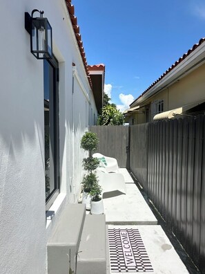 A narrow outdoor pathway leading to a building with a white exterior, a metal fence, and a welcome mat. - Casita Near Little Havana with Parking (Miami)