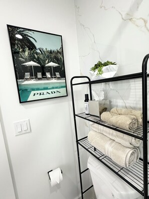 A bathroom with a black metal shelf holding towels and a bottle of shampoo, a framed picture of a pool, and a marble wall. - Casita Near Little Havana with Parking (Miami)