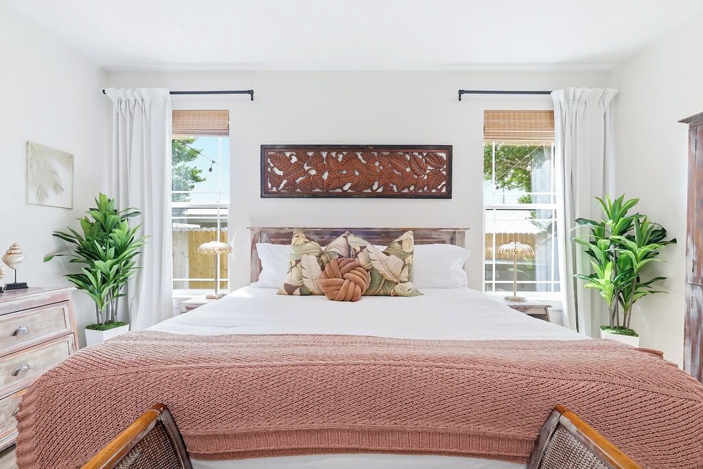 A bedroom with a large bed, a wooden headboard, and two windows with white curtains.