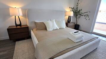 Un dormitorio con una cama, mesitas de noche, una lámpara, una planta en maceta y vistas a través de una ventana.