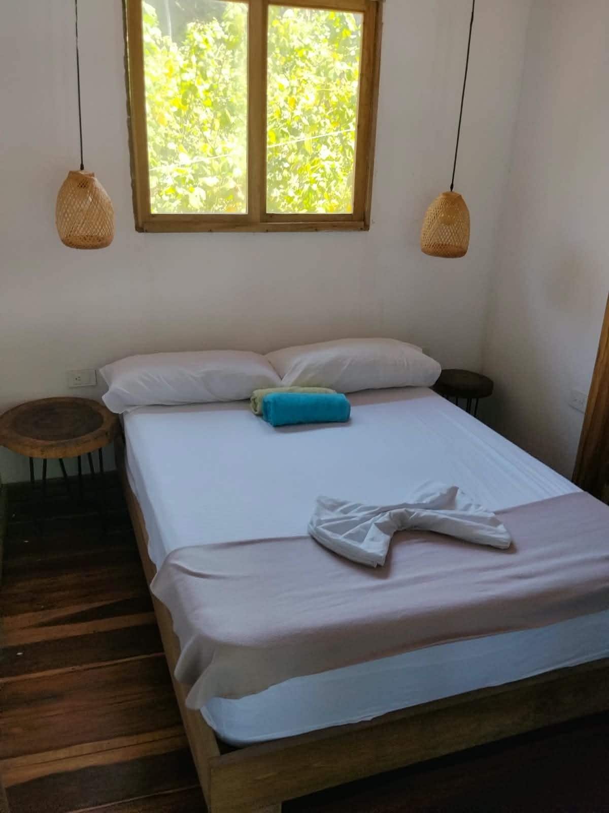 Uma cama de solteiro com lençóis brancos, um travesseiro azul e uma toalha branca sobre a cama. Há um banquinho de madeira redondo ao lado da cama.