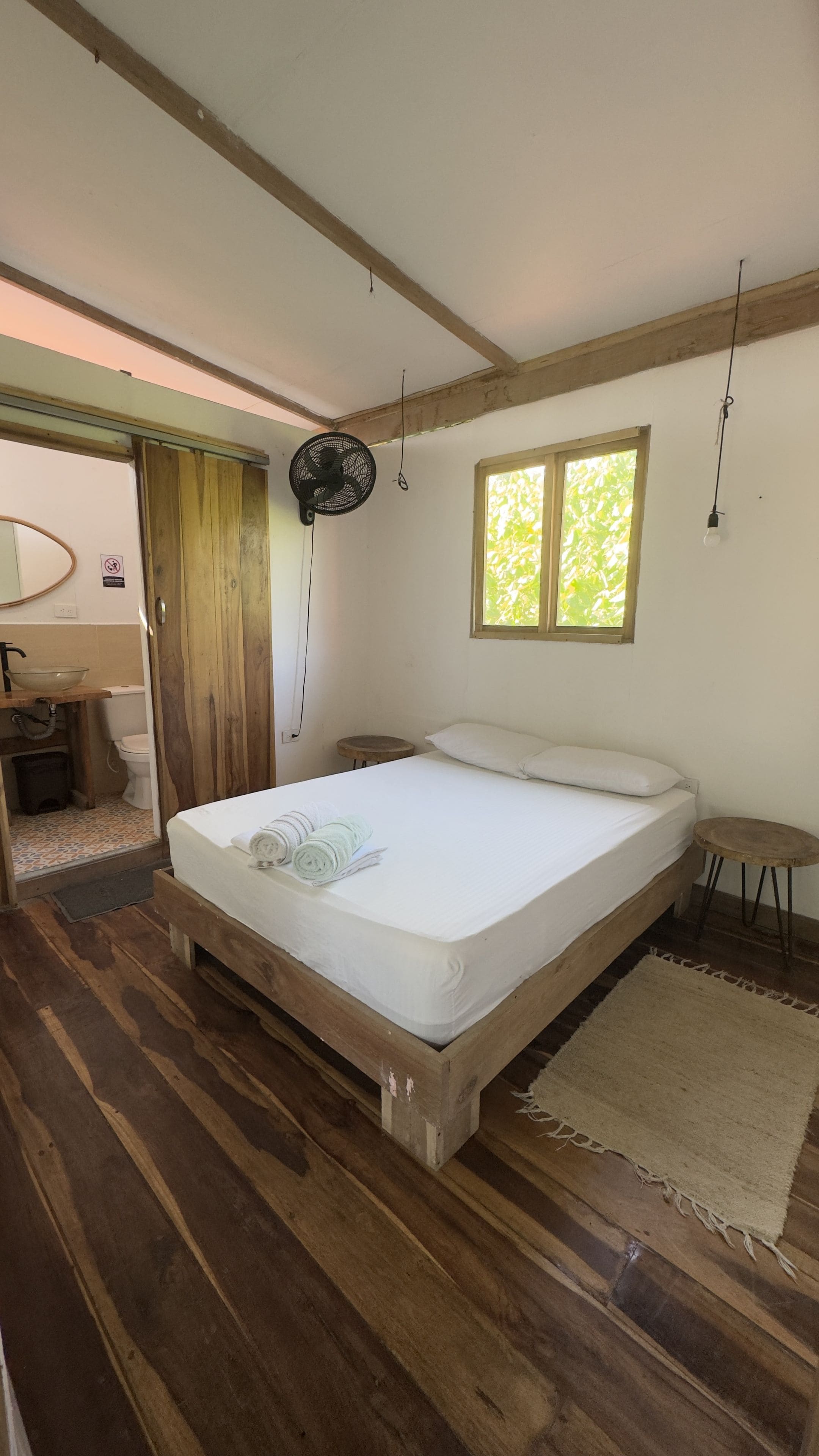 A bedroom with a wooden bed, a small bedside table, a window with natural light, and a fan on the ceiling.