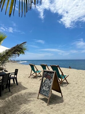 A beachside setting with a chalkboard sign advertising beers and water, outdoor seating, and a view of the ocean. - Eco Hostal Paraiso (Santa Marta)