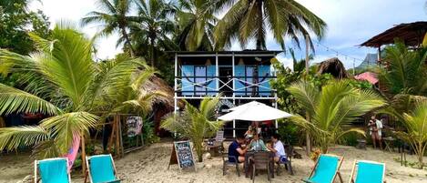 A beachside restaurant with outdoor seating, palm trees, and a building with a blue facade.