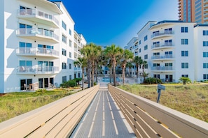 A modern multi-story residential building with balconies, a paved walkway, and palm trees. - Beachfront 2BR @ Palm Beach Resort • Sleeps 7 • Stunning Views (Orange Beach)