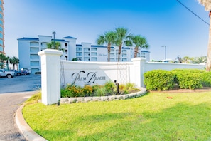 A hotel entrance with a sign reading 'Palm Beach,' surrounded by well-maintained grass and palm trees. - Beachfront 2BR @ Palm Beach Resort • Sleeps 7 • Stunning Views (Orange Beach)