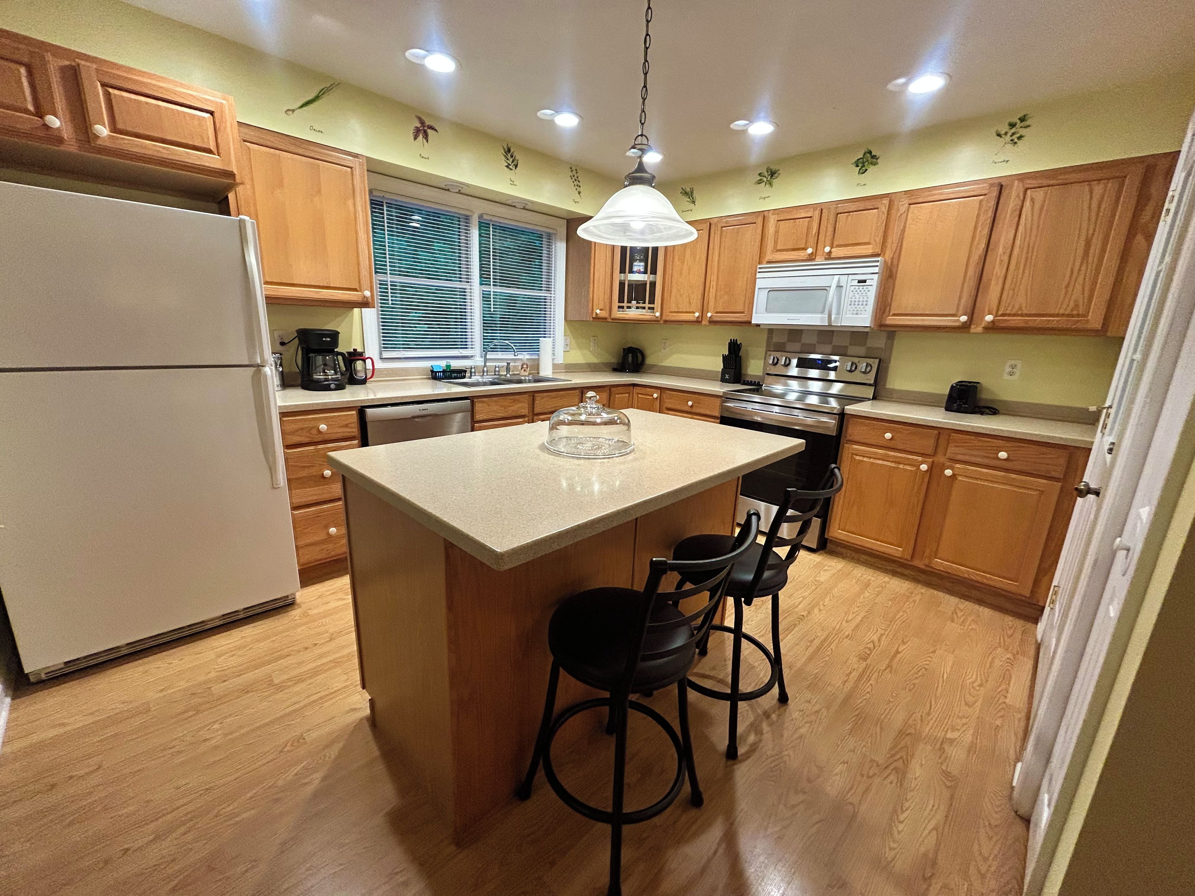 A kitchen with wooden cabinets, a central island with bar stools, a refrigerator, a stove, and a window with blinds.
