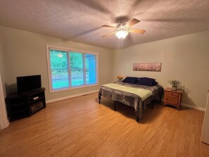 Quarto com cama, ventilador de teto, janela com persianas, rack para TV e mesa de cabeceira com planta.